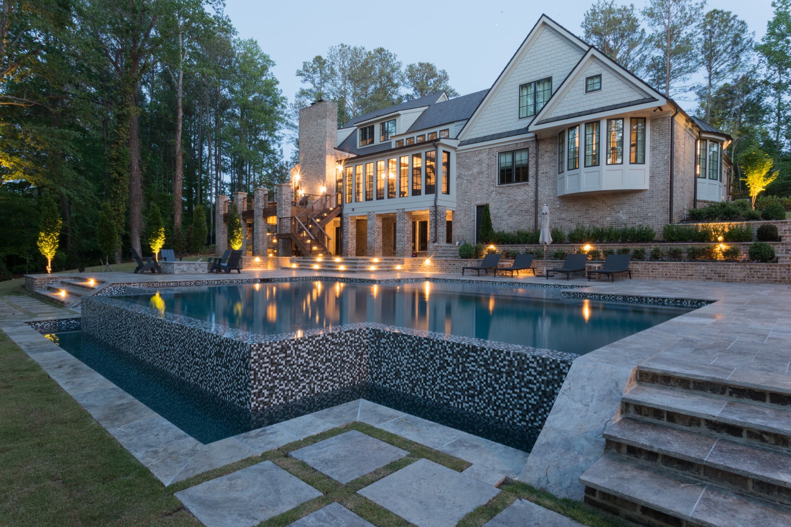 The Chadwell project in Roswell at twilight: a linear modern pool with a vanishing edge and glass-tile spillway below a brick estate