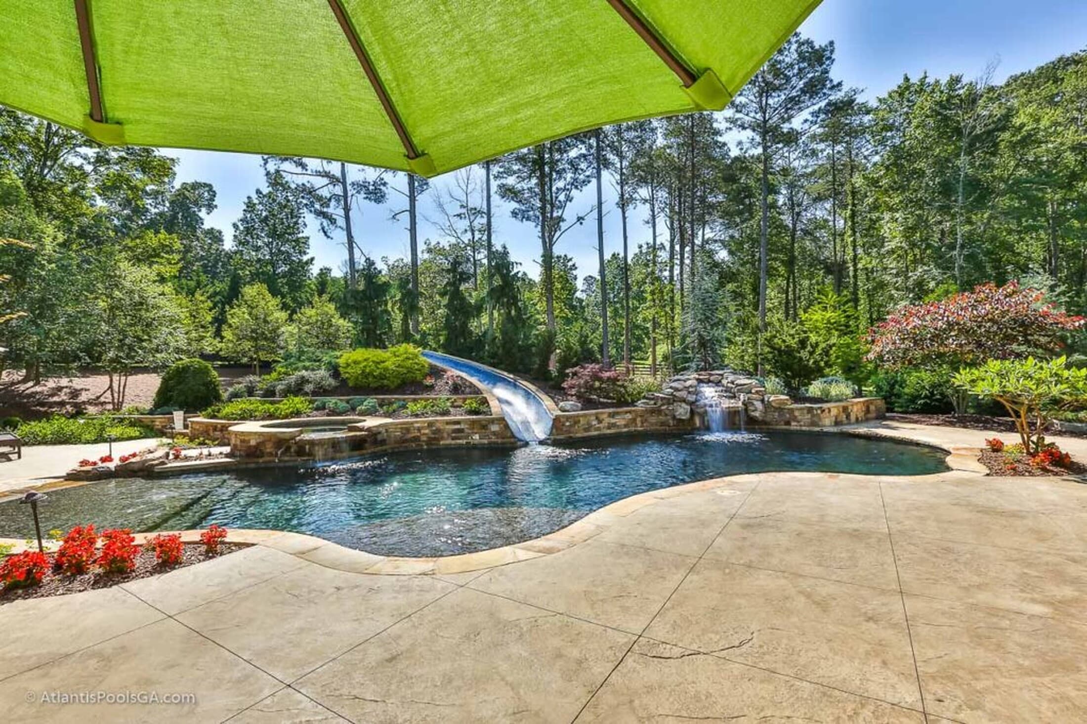 A wide freeform pool with a water slide, waterfall, and mature woodland beyond