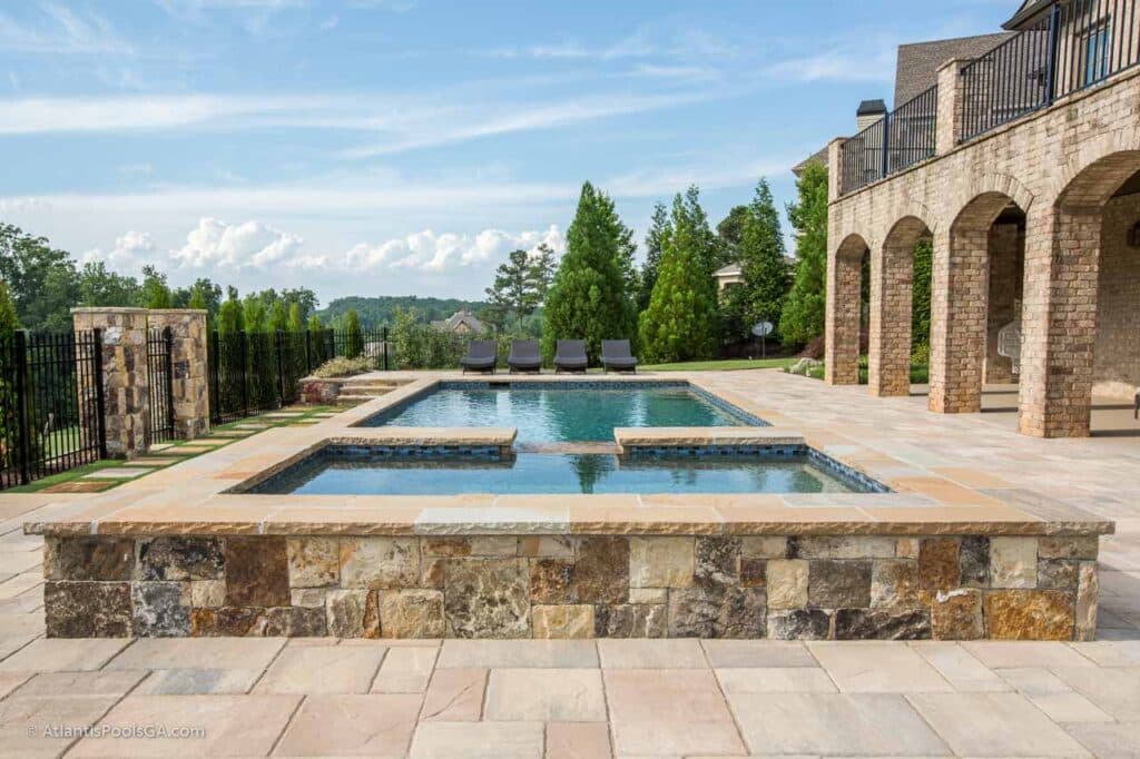 A rectangular pool and raised spa framed by a stone deck, planted evergreens, and a brick home with arches.