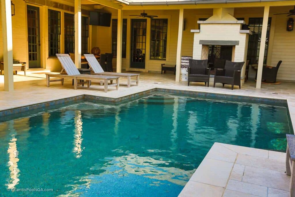 A rectangular linear pool set against a covered patio with a stone fireplace, the straight pool edge running parallel to the house
